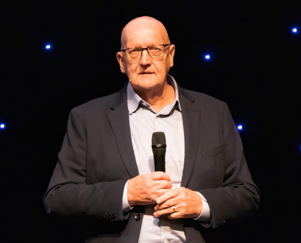 Martin Evan's in a suit holds a microphone on stage at the Catford Theatre, London with a black background featuring small illuminated spots.