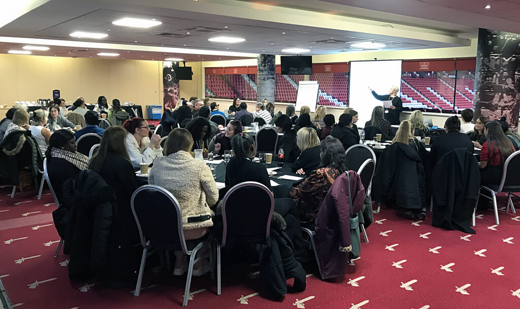 A large group of people seated at tables in a conference room watching Caroline Braxton of The Training Network in front of a screen.