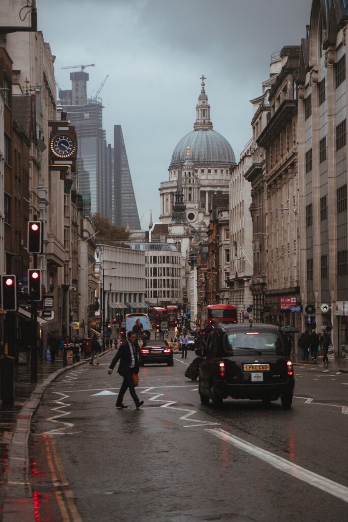 A rainy city street with a dome-topped cathedral in the background is surrounded by tall buildings. Amidst the bustle of black cabs and red buses, doctors hurrying to provide primary care walk alongside others with umbrellas on the slick pavement.