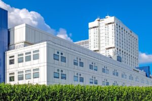 A modern white hospital building with multiple windows stands in front of a taller white skyscraper, under a blue sky with clouds, partially obscured by a green hedge.