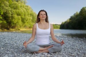 A woman sits cross-legged on a rocky riverbank, meditating with her eyes closed. She wears a white top and gray pants, much like medical staff in moments of calm between responsibilities, surrounded by greenery and the soothing flow of the river.