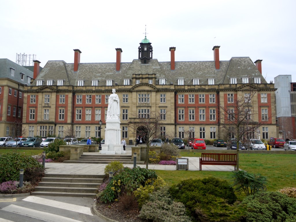 The Royal Victoria Infirmary is a large historic building with a central tower, numerous chimneys, and a statue in front. It stands proudly amidst parked cars and landscaped gardens beneath a cloudy sky, embodying the grandeur and service of the NHS.