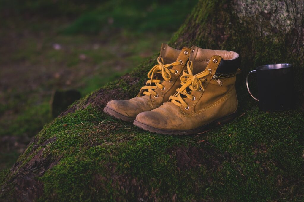 A pair of well-loved yellow hiking boots that fit perfectly, featuring yellow laces, rest on a moss-covered tree base beside a black metal mug.