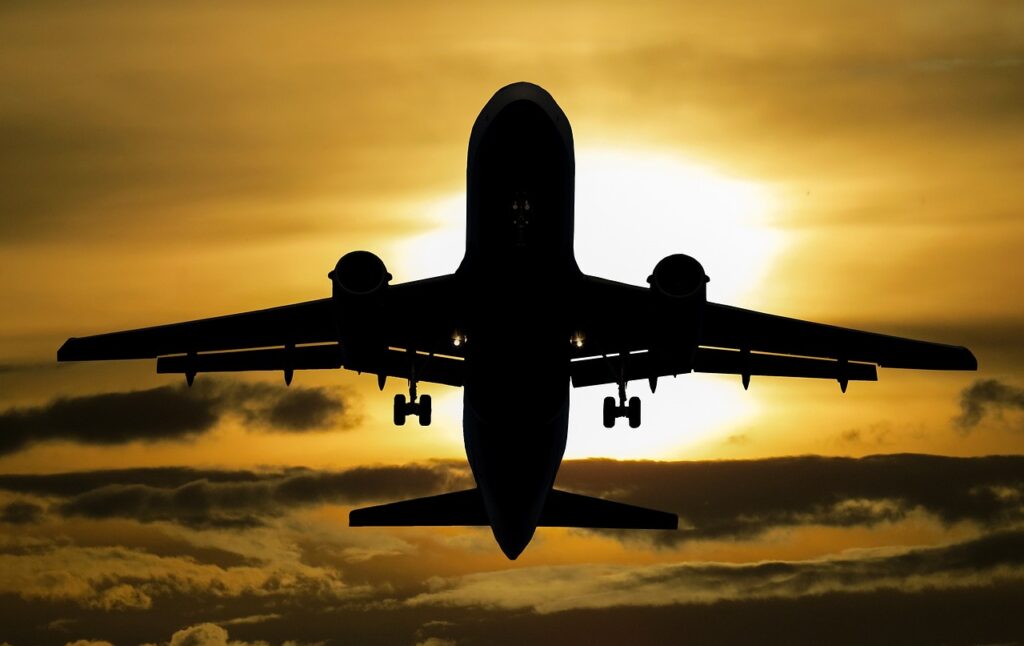 A silhouette of an airplane in flight against a bright yellow and orange sunset sky with scattered clouds.