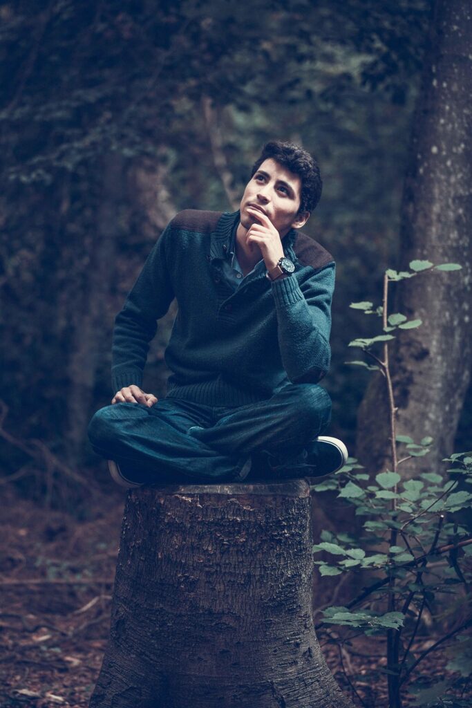 A man sits cross-legged on a tree stump in a forest, looking upward with one hand resting on his chin. He wears a blue sweater, jeans, and a watch.