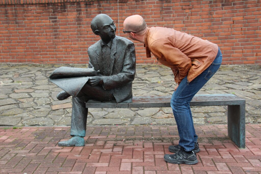 A man in a brown jacket leans toward a seated bronze statue of a man holding an NHS newspaper on a bench, with a brick wall in the background.