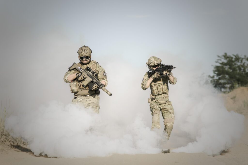 Two soldiers in camouflage uniforms and tactical gear walk through a cloud of smoke outdoors, holding rifles in a training or combat scenario.