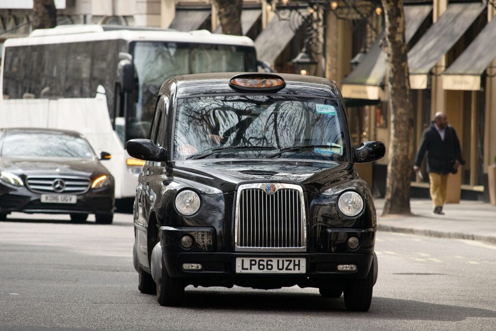 raditional London black cab driving through a busy city street with shops and traffic in the background.