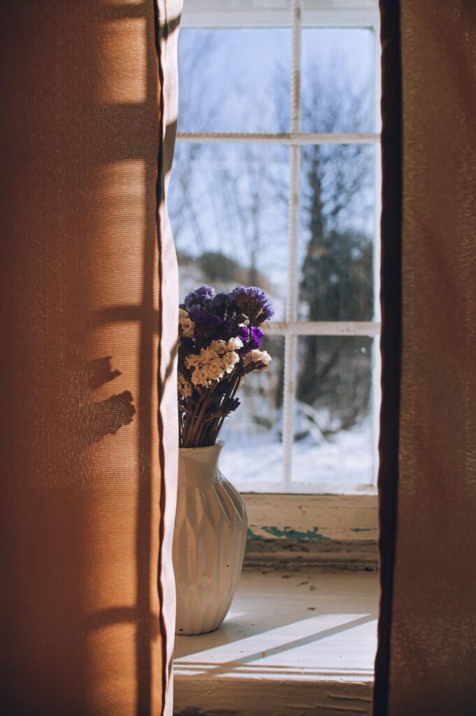 A vase of purple and white dried flowers sits on a sunlit windowsill. The window looks out onto a snowy, blurred landscape with trees. Brown curtains are pulled back on either side.