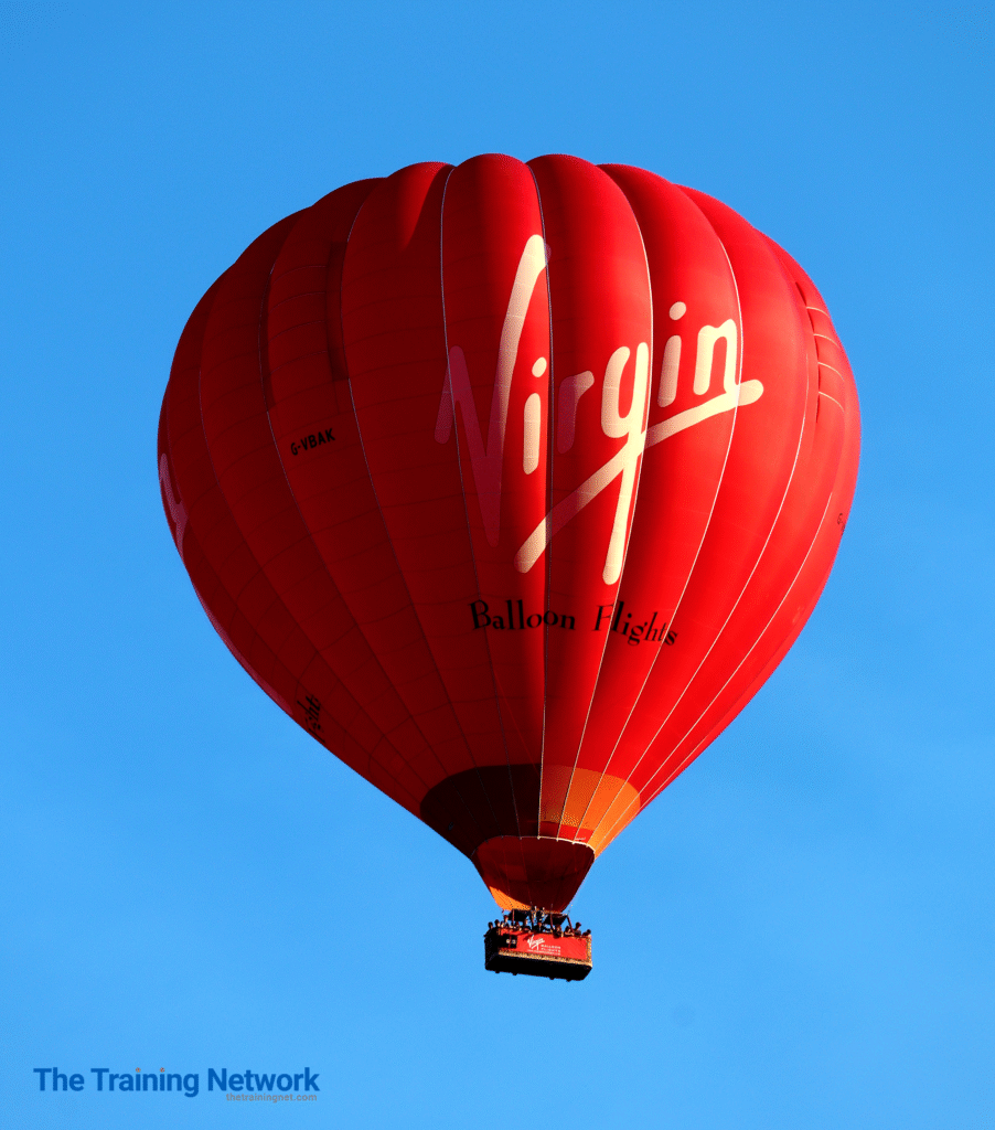 A large red hot air balloon with the word "Virgin" written in white cursive, and "Balloon Flights" underneath it, floats against a clear blue sky.