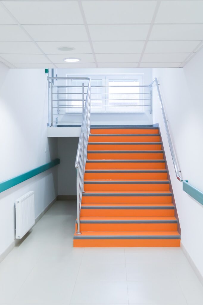 Empty hospital stairwell with bright orange steps, suggesting institutional transition and the risks of creating bottlenecks through poorly sequenced organisational change.