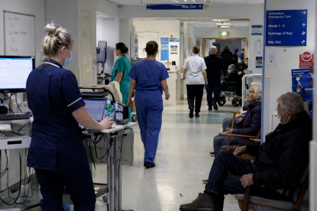 A busy NHS hospital corridor with nurses and doctors moving between wards, while a small number of patients sit waiting on chairs along the wall under bright clinical lighting.