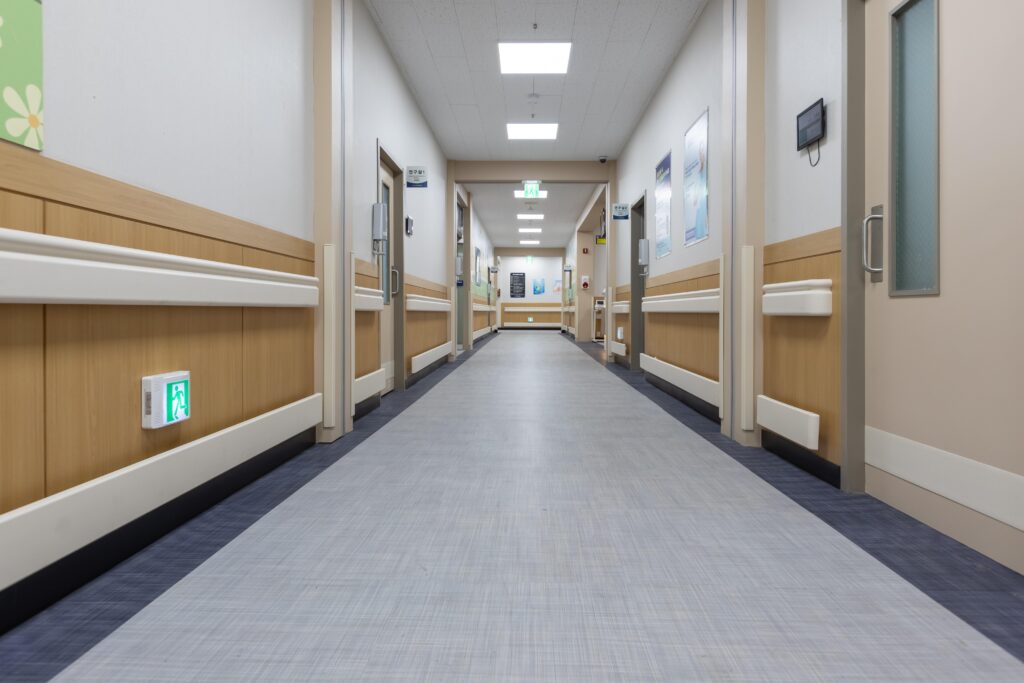 Empty hospital corridor with clean floors, handrails, and patient room doors on both sides, brightly lit with ceiling lights