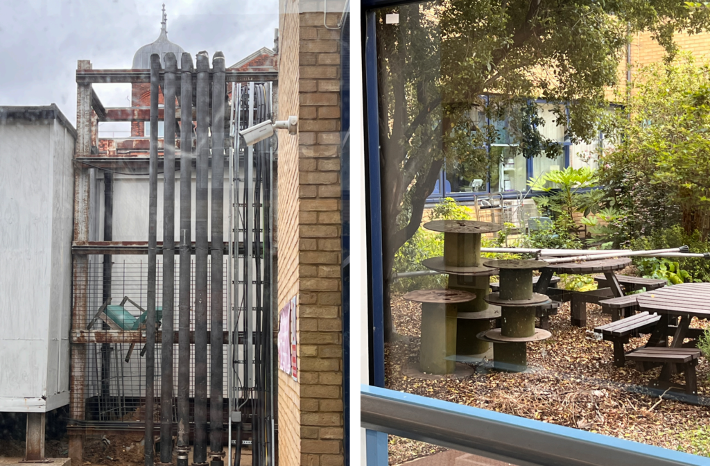 View from inside a hospital building looking through windows onto neglected service areas and courtyards. One side shows rusting industrial pipework, metal framework and a security camera between buildings. The other shows discarded wooden cable reels repurposed as tables among overgrown planting, fallen leaves and outdoor seating, creating a contrast between institutional infrastructure and improvised outdoor space. NHS patient experience and hospital leadership