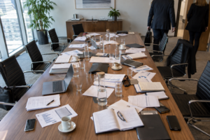 Overhead view of a senior management meeting room after a meeting, with a long table covered in briefing papers, notebooks, laptops, water glasses and coffee cups. Empty chairs surround the table while two officials leave the room in the background. NHS England abolition feasibility