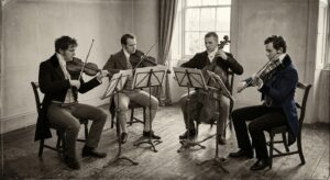 Four classical musicians performing a string quartet in a nineteenth-century rehearsal room, illustrating Baumol’s cost disease and the limits of NHS productivity. NHS productivity and patient pathways
